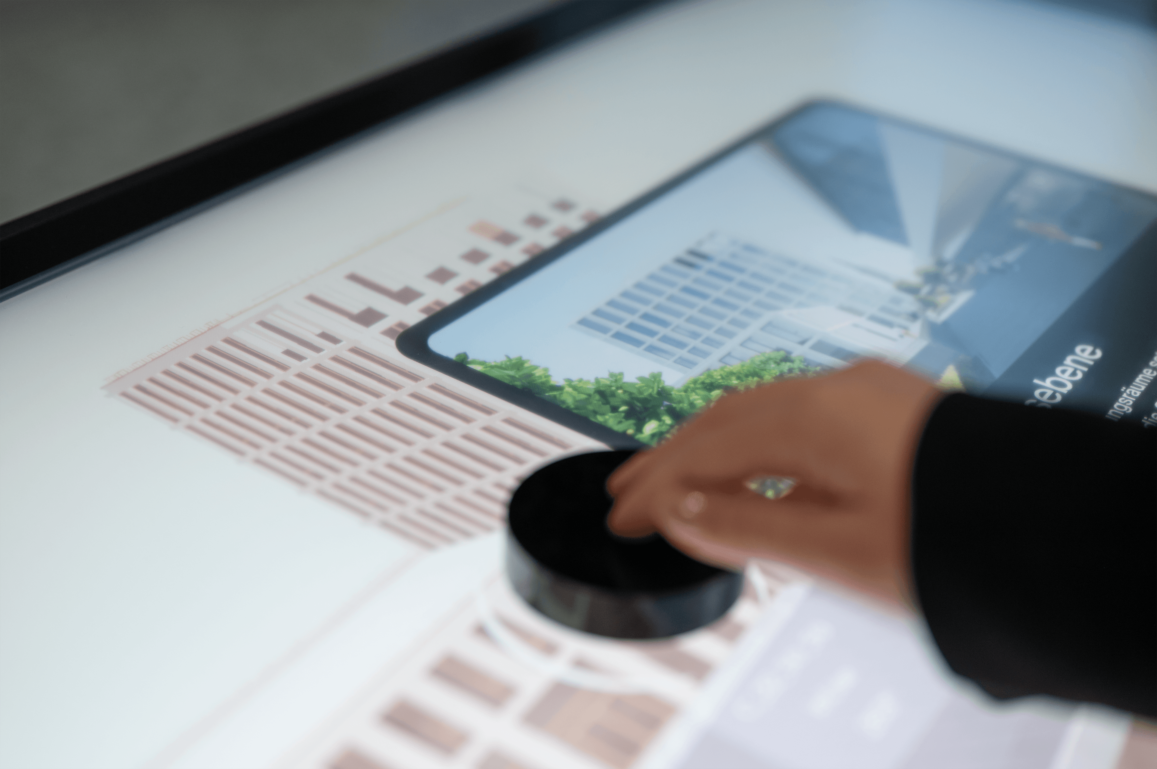 Eine Hand bewegt einen schwarzen Puck auf dem Tisch, auf dem ein 3D-Architekturmodell des SGP-Campus in Ulm und ein besonderer Bereich darauf zu sehen sind.