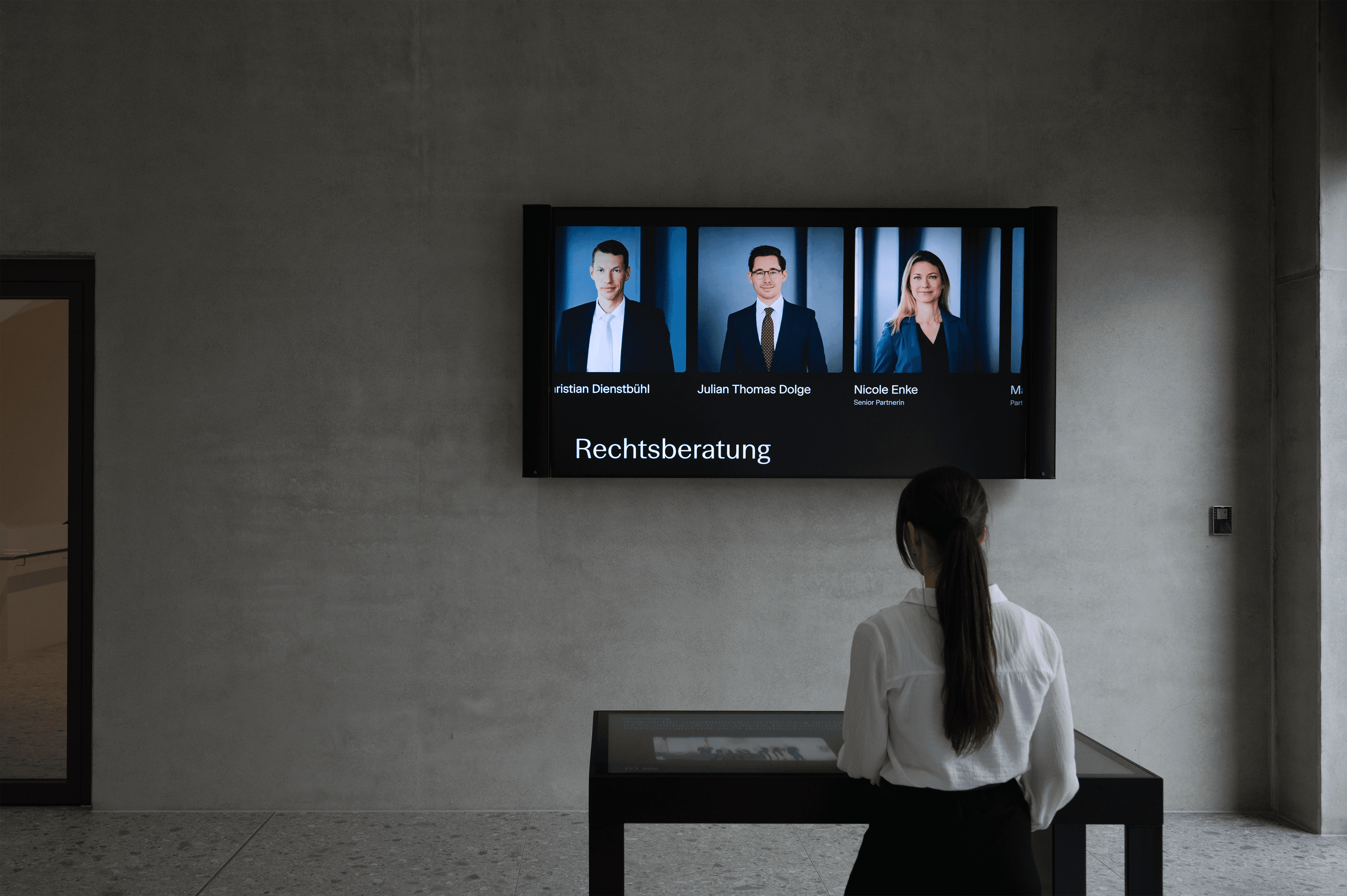 Eine Frau betrachtet in einem modernen, minimalistischen Raum einen an der Wand montierten Bildschirm, auf dem „Rechtsberatung“ und drei Porträtbilder zu sehen sind.