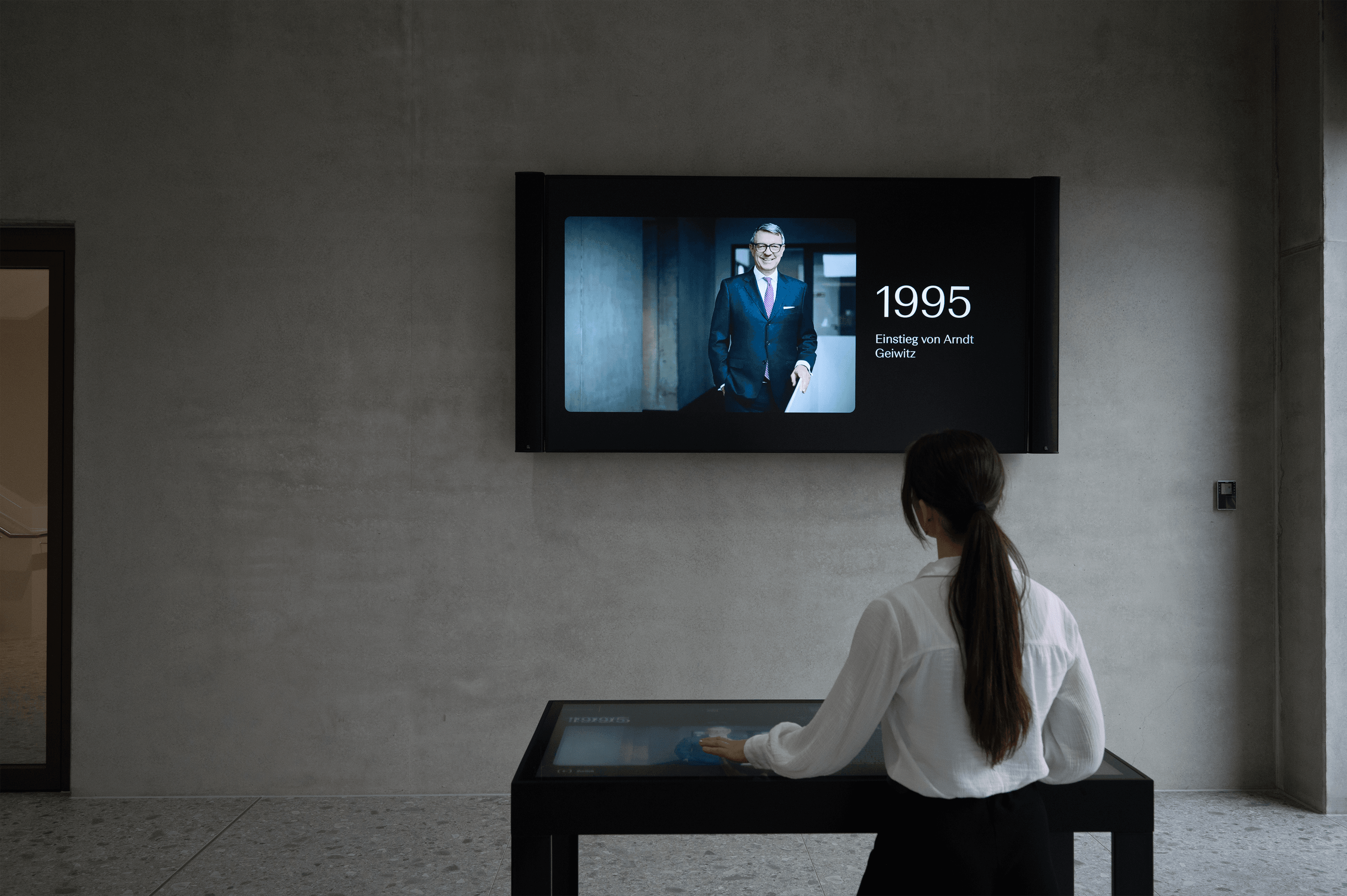Eine Frau berührt einen interaktiven Tisch, während sie auf einen großen Wandbildschirm blickt, auf dem das Porträt von Arndt Geiwitz zu sehen ist.