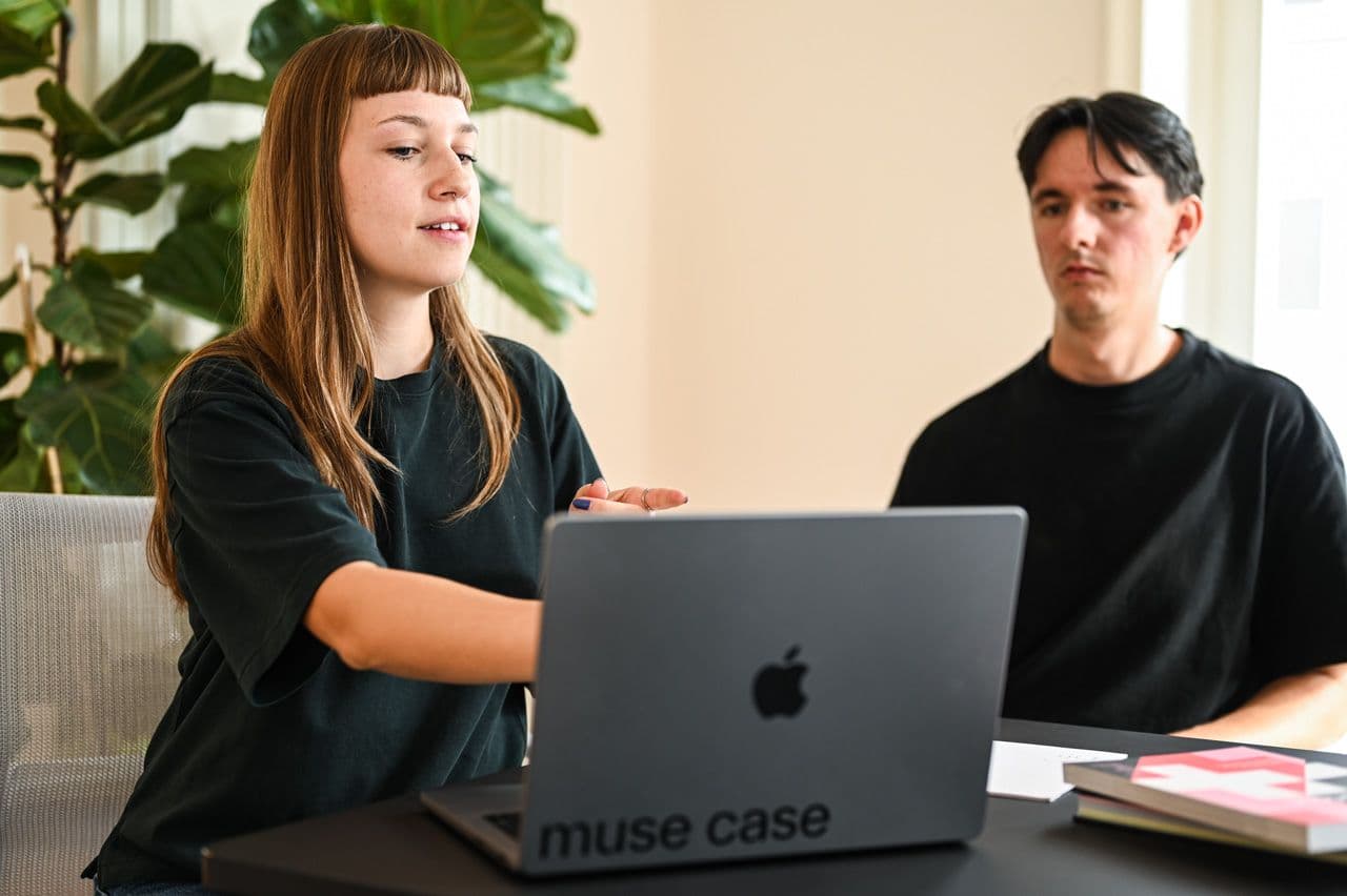 Zwei Personen in einem Meeting, eine Frau spricht und gestikuliert, während ein Mann zuhört. Vor ihnen steht ein muse case MacBook auf einem Tisch mit Unterlagen.