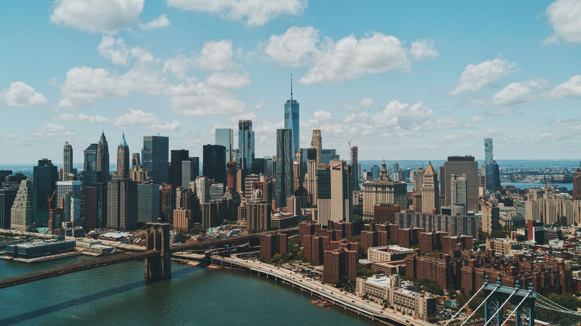 Aerial view of the New York City skyline featuring One World Trade Center, the Brooklyn Bridge, and the East River.
