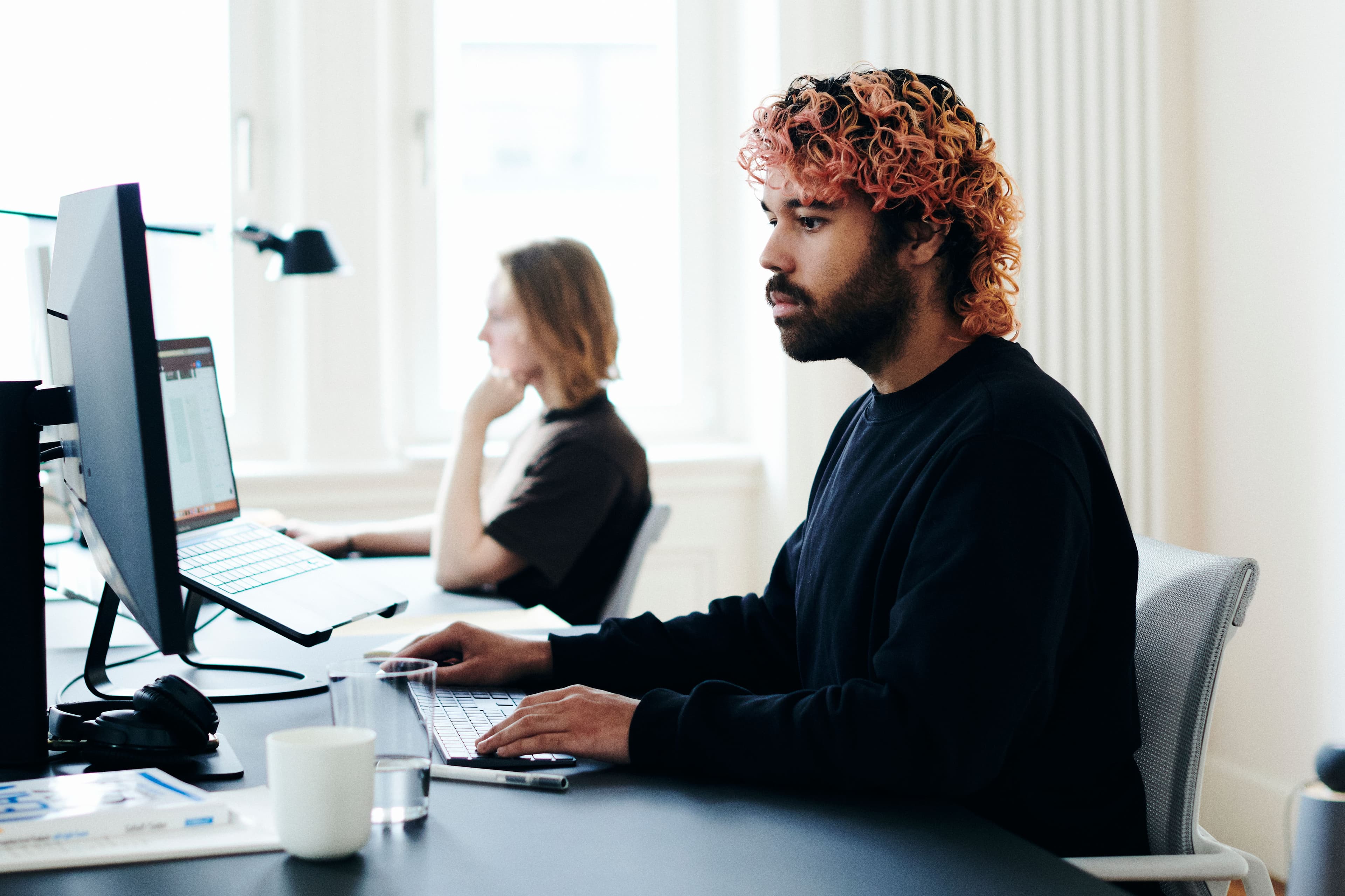 Person mit lockigem, rosa gefärbtem Haar arbeitet an einem Desktop-Computer in einem hellen Büro, während im Hintergrund eine weitere Person an einem Laptop sitzt.